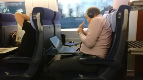 Mann schaut im ICE Film auf dem Tablet, während er isst und sein Arbeits-Notebook vor ihm steht