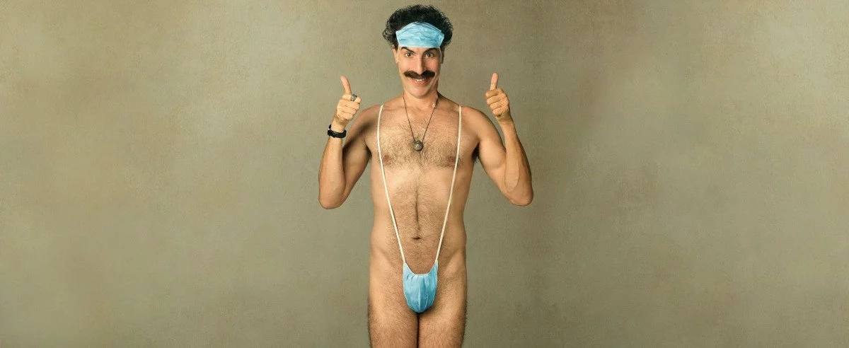 Nackter Borat zeigt Daumen hoch mit Maske auf Stirn und im Schrittbereich