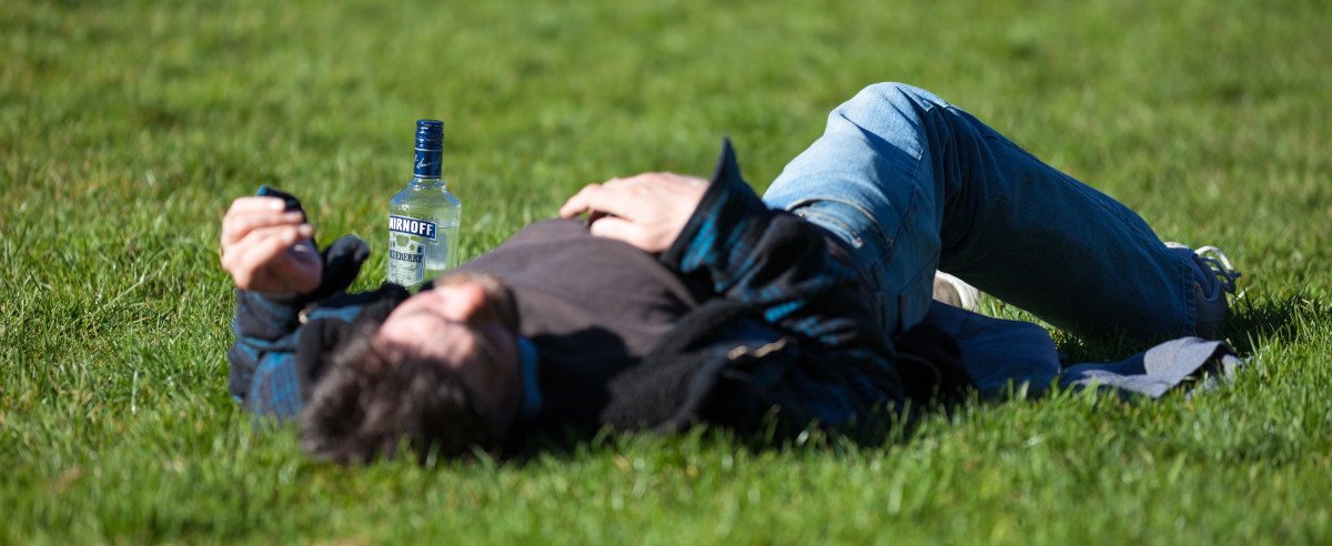 Ein Mann, der betrunken auf einer Wiese liegt, neben ihm eine Flasche Wodka.