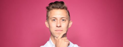 Ein junger, weißer Mann steht vor einem farbigen Hintergrund und greift sich fragend ins Gesicht