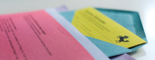 Ein pinkes Begleitschreiben liegt auf einem blauen Umschlag, in dem ein gelber Stimmzettel steckt