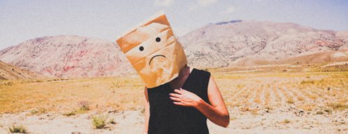 Mensch steht in der Wüste und hat eine Papiertüte über den Kopf, auf der ein trauriger Emoji gemalt ist