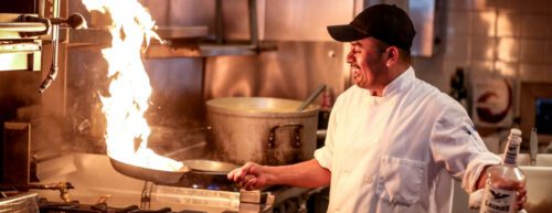Ein Koch hält eine Pfanne in der Hand, aus der Flammen züngeln