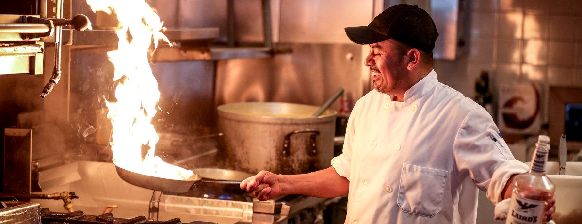 Ein Koch hält eine Pfanne in der Hand, aus der Flammen züngeln